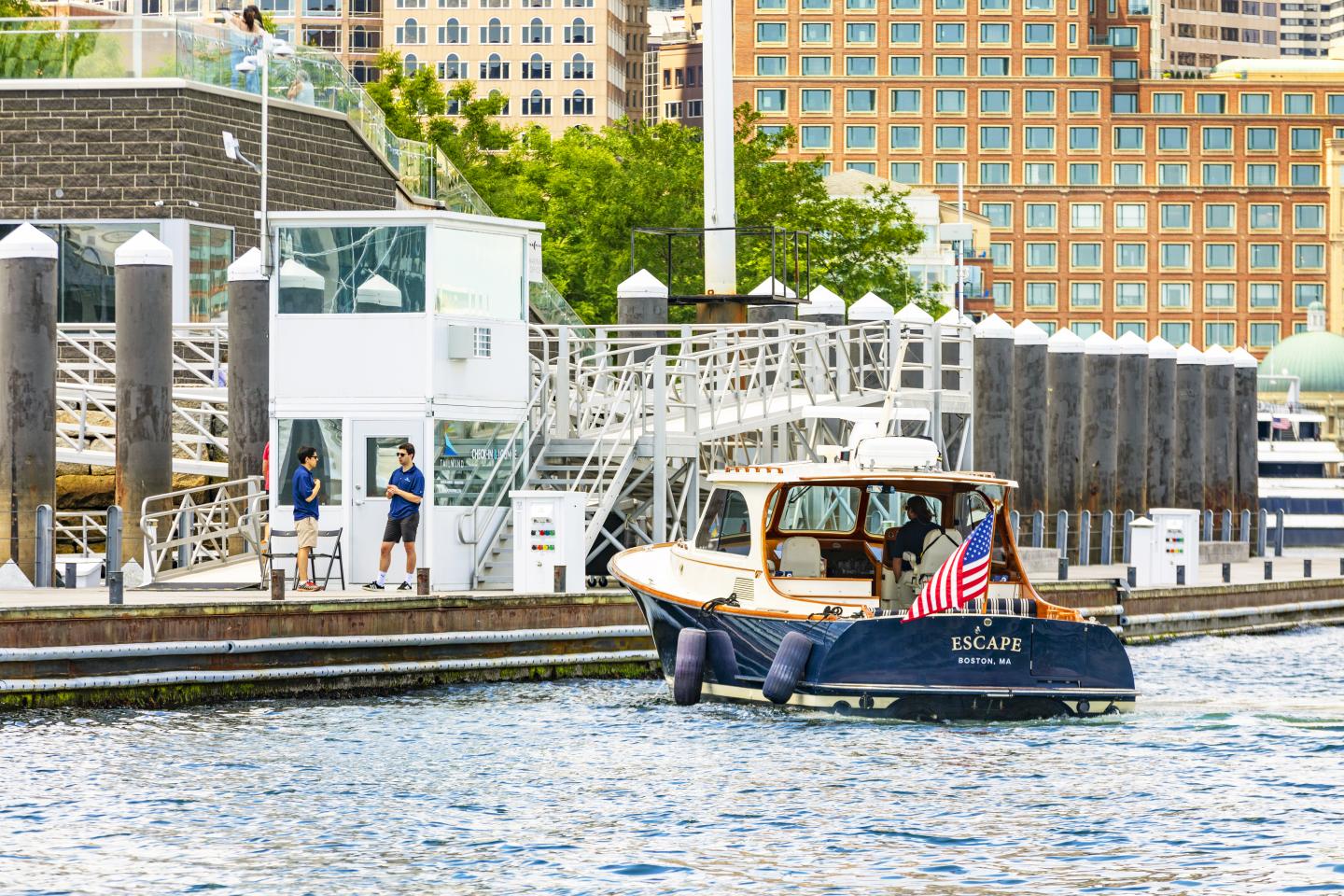 Boat with an American flag near a dock, city buildings in the background.